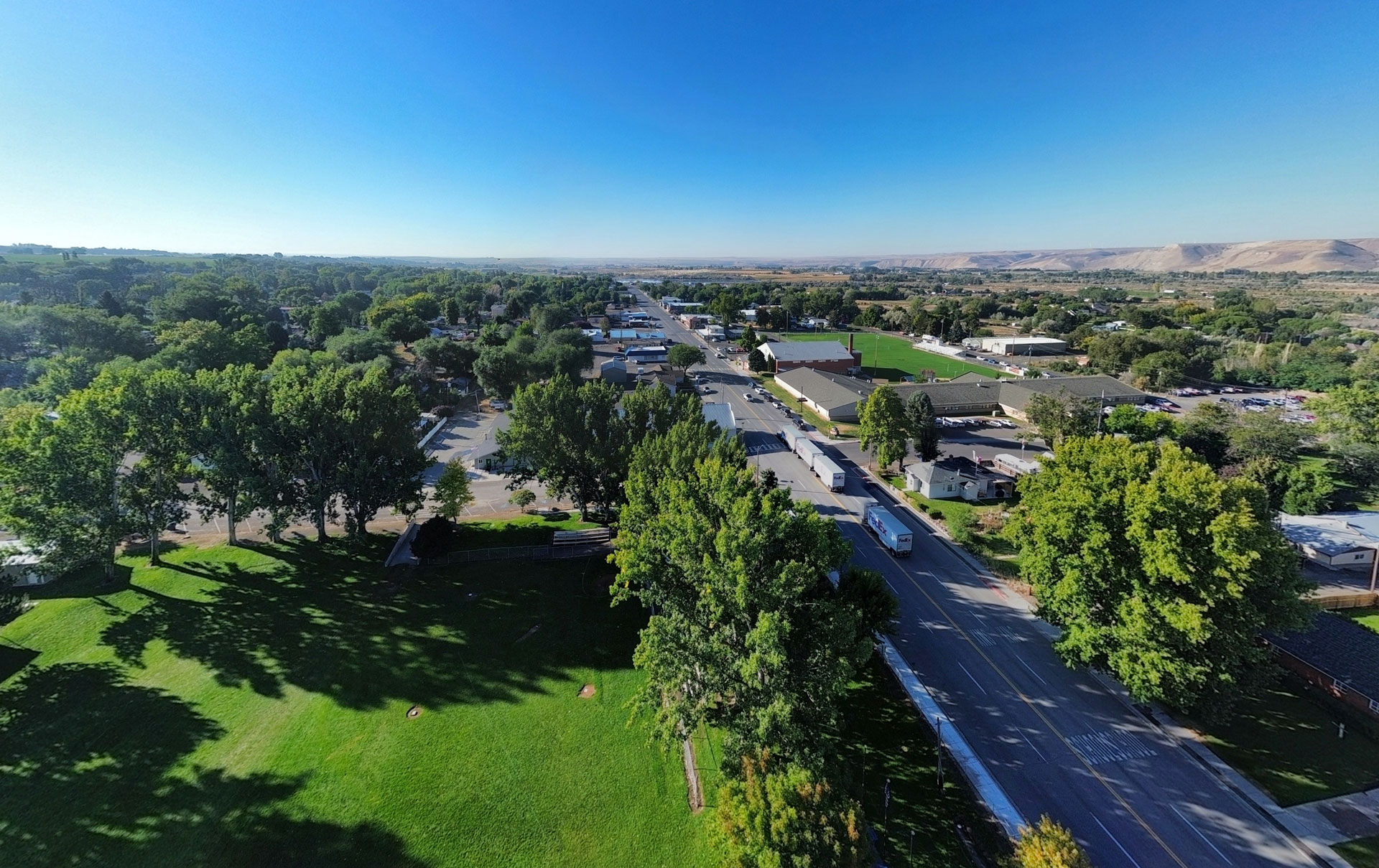 Aerial view of a small town with tree-lined streets, green parks, and buildings under a clear blue sky, with distant hills visible on the horizon. Hagerman, ID