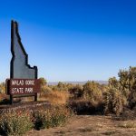 A sign shaped like the state of Idaho reads Malad Gorge State Park, surrounded by dry bushes and grass under a clear blue sky. Hagerman, ID