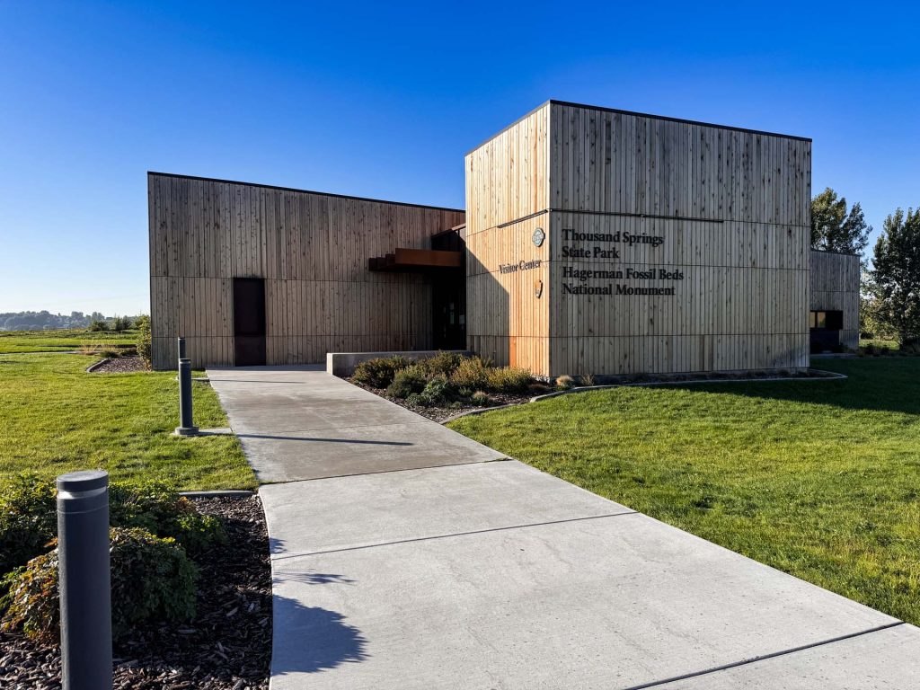 A modern wooden building with signs for Thousand Springs State Park and Hagerman Fossil Beds National Monument, set on a landscaped lawn under a clear blue sky. A paved walkway leads to the entrance. Hagerman, ID