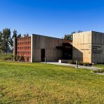 Modern visitor center building with wooden exterior surrounded by green grass and trees, under a clear blue sky. Signs on the building indicate it is for a national monument. Hagerman, ID