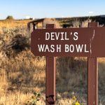 A brown wooden sign with white letters reads “DEVIL’S WASH BOWL,” standing in a dry, grassy field under a clear sky. Shrubs and distant structures are visible in the background. Hagerman, ID