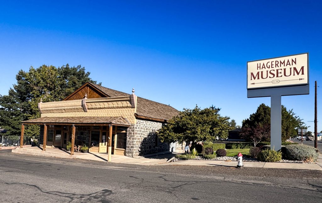 A small stone building with a wooden porch sits by a road under a clear blue sky. A large sign reads Hagerman Museum. Trees and shrubs surround the museum, and a fire hydrant is visible in front. Hagerman, ID