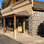 A historic stone and wood building with a covered porch and large windows sits on a quiet street. Signs are posted on the door, and a bench and potted plant are visible by the entrance. Hagerman, ID