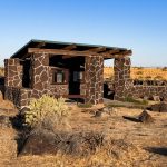 A small, open stone shelter with a green roof stands in a dry, desert landscape with brown grasses and shrubs under a clear blue sky. The structure features dark, irregular stone walls and wooden beams. Hagerman, ID