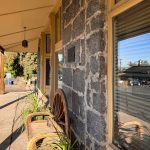A stone building with a bench and wagon wheel outside, plants nearby, and large windows reflecting parked cars and trees. The sidewalk runs alongside a quiet street on a sunny day. Hagerman, ID