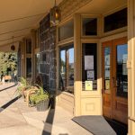 A sunlit building entrance with large windows, a wooden door, potted plants, and flyers posted on the glass. The sidewalk and part of the street are visible, with shadows cast by the overhang. Hagerman, ID