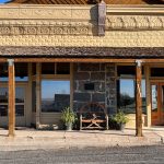 Historic storefront with tan decorative brickwork, wooden beams, and large windows. A stone wall section holds a bench made from a wooden wagon wheel, flanked by potted plants. Sunlight casts shadows on the sidewalk. Hagerman, ID