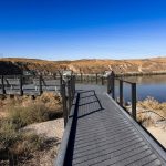 A wooden boardwalk with railings leads to an overlook platform above a wide river, surrounded by dry, brown hills under a clear blue sky. Informational signs are visible on the platform. Hagerman, ID