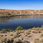 A calm river bordered by dry, scrubby vegetation and rocky hills under a clear blue sky, with faint power lines overhead. Hagerman, ID