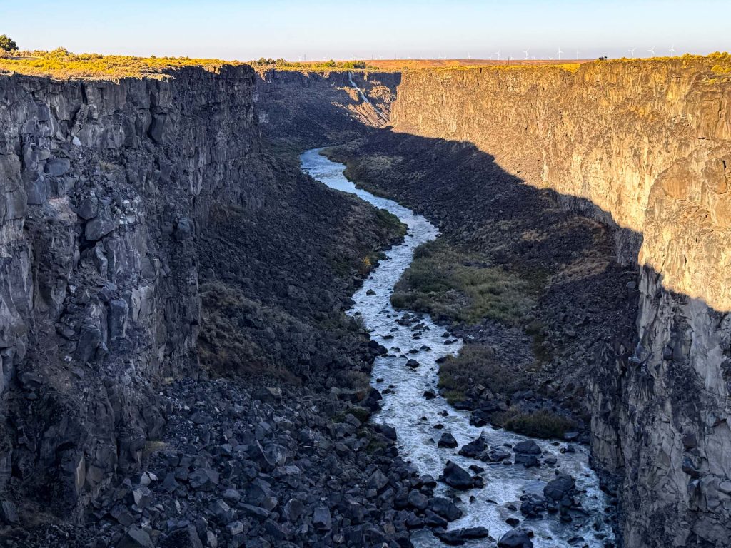 A narrow river flows between steep, rocky cliffs in a deep canyon under sunlight, with dry grassland visible on top of the cliffs and blue sky in the background. Hagerman, ID