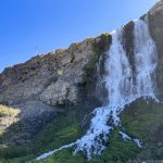 A waterfall cascades down a rocky cliffside, surrounded by green vegetation at the base, under a clear blue sky with sunlight illuminating the scene. Hagerman, ID