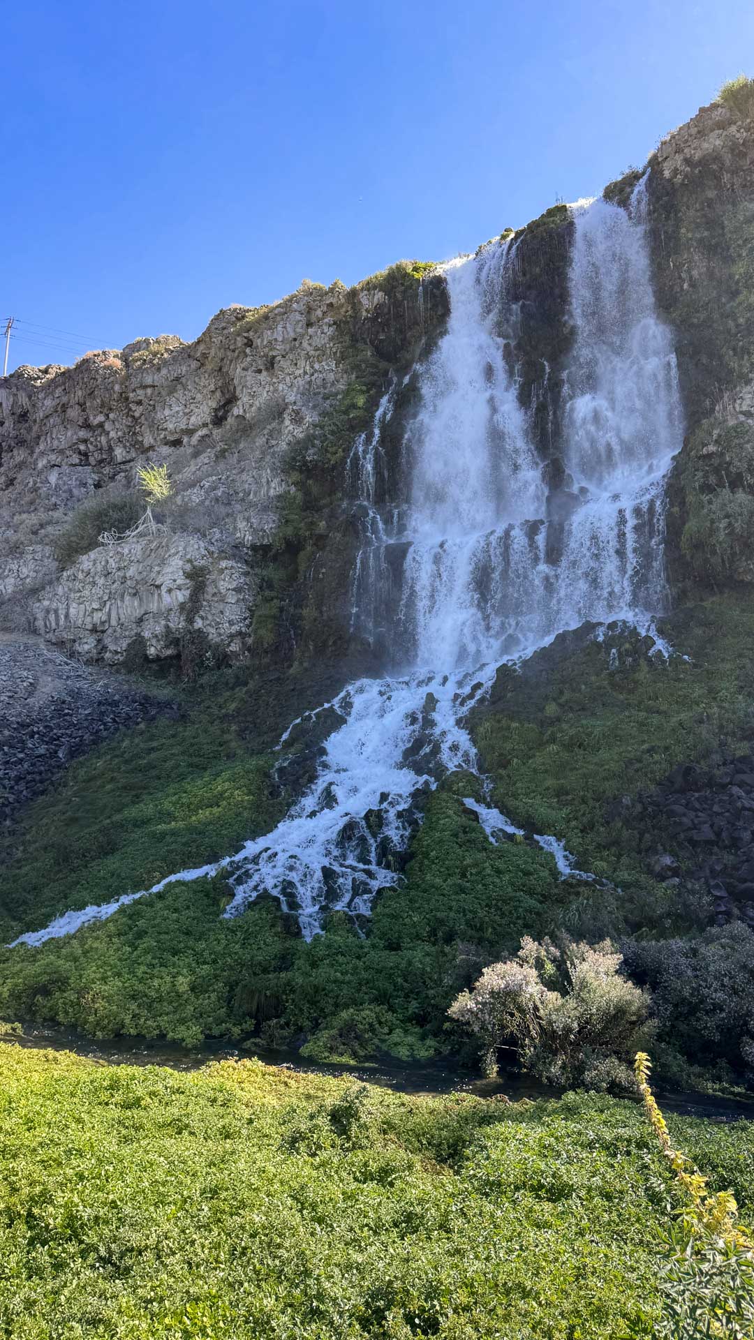A large waterfall cascades down a rocky cliff covered in greenery under a clear blue sky, with lush vegetation at the base of the falls. Hagerman, ID