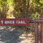 A wooden sign reading River Trail with a white arrow points forward at the entrance to a shaded dirt path surrounded by trees and sunlight. Hagerman, ID