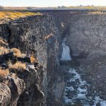 A waterfall flows into a rocky canyon with steep cliffs, while a bridge spans the top of the canyon. Dry grass and shrubs grow along the cliff edges under a clear sky. Hagerman, ID