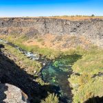 A panoramic view of a rocky canyon with steep cliffs and a winding river below, surrounded by green vegetation and sparse brush under a clear blue sky. Hagerman, ID