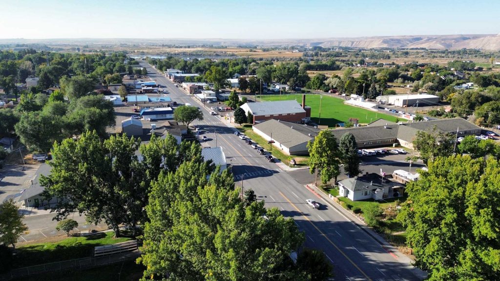 Aerial view of a small town with wide streets, green trees, buildings, and open fields; hills and farmland can be seen in the background under a clear sky. Hagerman, ID