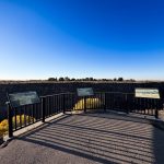 A viewing platform with three informational signs overlooks a deep canyon under a clear blue sky; shadows from the railing stretch across the paved ground. Sparse vegetation and distant trees are visible in the background. Hagerman, ID