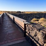 A metal footbridge leads across a rocky ravine with dry grass and bushes, under a clear blue sky. A small stone building appears in the distance on the other side of the bridge. Hagerman, ID